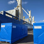 Blue-wrapped industrial equipment staged on a quay beside a geared cargo vessel during project cargo handling operations at the Port of Hamina.